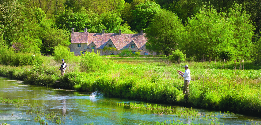 Landscape photo of two men fishing in a river in front of a Cotswolds barn